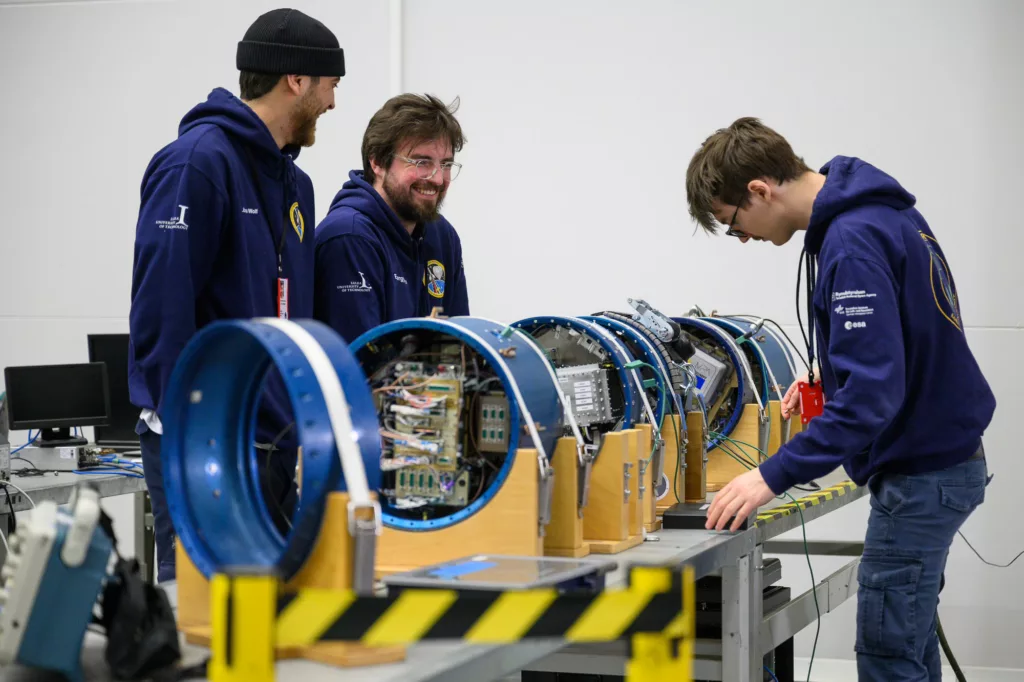 Three space students prepare science rocket modules placed together on a table.