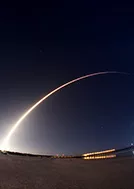 A long-exposure photog of a rocket launch. The rocket's trajectory is depicted as a bright, curved streak of light against the dark night sky.