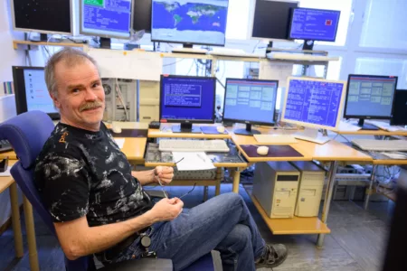 A man sitting back in his office chair in front of nine computer screens for the Odin satellite, smiling at the camera